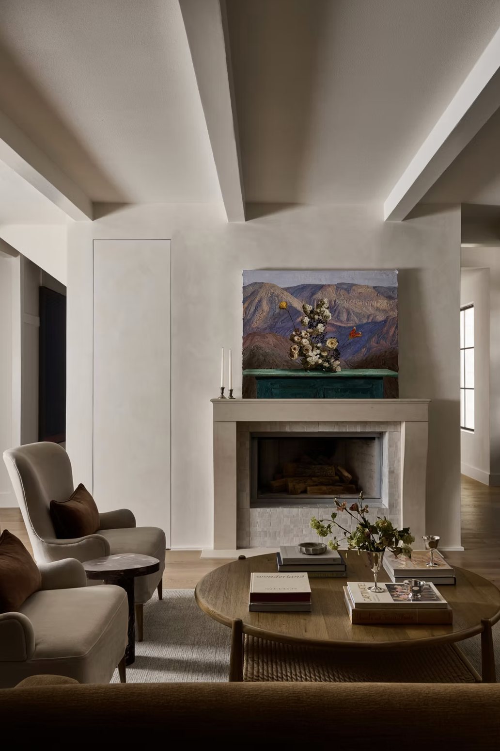 Limestone fireplace on a plaster wall beneath painted beams, with a landscape painting on the mantel