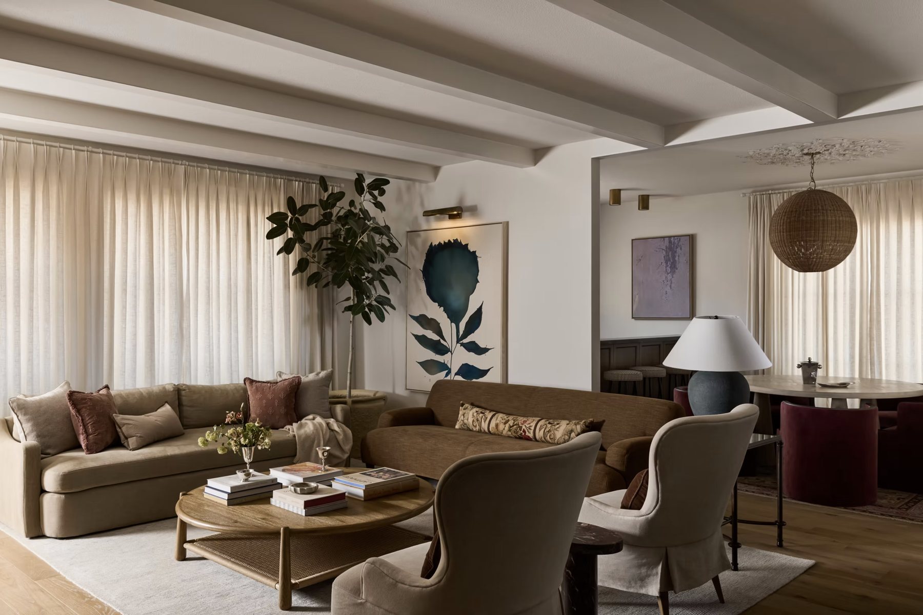 Living room under painted beams with linen drapery, curved sofas and wingback chairs, opening to the dining room