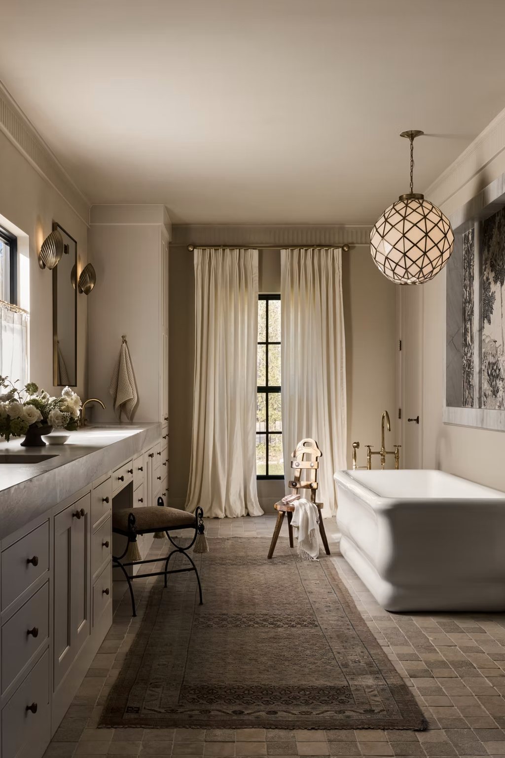 Primary bath with a long stone vanity, freestanding tub, linen drapery and a faceted glass pendant